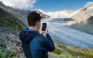 Young man with a backpack and hiking equipment walks in the mountains and takes a photo on the mobile phone of a beautiful nature.Traveler enjoys the scenery and shares photos.