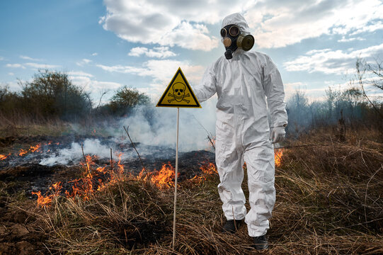 Fireman ecologist fighting fire in field. Man in protective radiation suit and gas mask near burning grass with smoke, holding yellow triangle with skull and crossbones warning sign.
