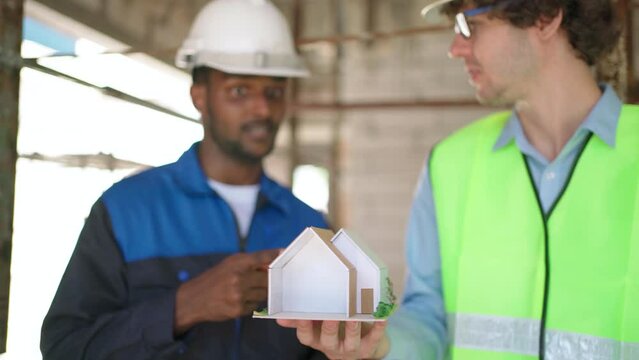 Architect and civil engineer in safety uniform holding house model structure and discuss at construction site on sunny day. Renovate and construction concept. Specialists home project