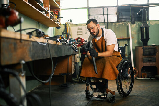 A manufacturer in a wheelchair grinds a metal part in his workshop.