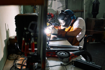 A metalworker in a wheelchair welds a metal parts in a workshop.