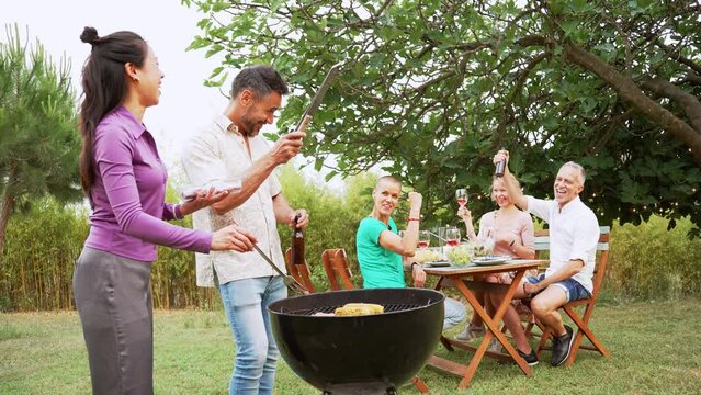 Couple cooking on a barbaque in countryside. At the back, his friends celebrating with alcohol drinks.