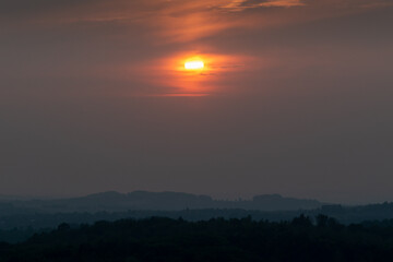 Dramatic apocalyptic sinister scene of bright orange sun behind clouds over silhouette forest layers