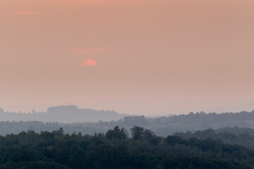 Atmospheric landscape scene of lush forest layers in mist during autumn sunset in countryside, forest obscured in fog and big orange sun dimmed behind clouds