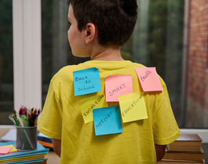 Rear view of a multitasking primary school student with colored paper for notes glued on his bright yellow t-shirt with inscriptions: Back to school, Smart Motivation Knowledge Erudition Education