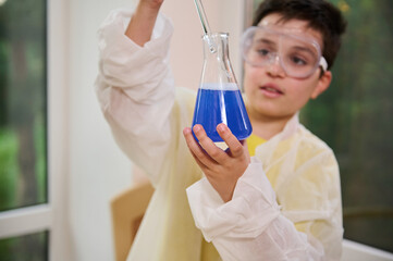 Focus on flat bottomed flask with chemical liquid in the hand of schoolboy chemist scientist wearing protective clothes, using glass stick to mix reagent during experiment in school chemistry lab.