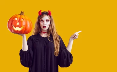 Schilderijen op glas Portrait young woman in Halloween costume standing isolated on yellow background, holding Jack o lantern, pointing finger at copyspace side and looking at camera with funny surprised face expression © Studio Romantic