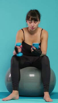 Vertical video: Bored woman using smartphone and trying to do workout exercise with dumbbell in hand, sitting on fitness toning ball. Person feeling tired after physical training with sport equipment.