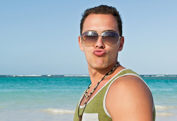 The man has pursed his lips and is fooling around. Portrait of young adult man in sunglasses against turquoise water in the sea on beach