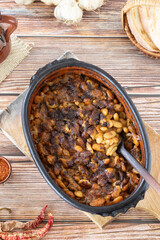 Spicy baked bean stew in a clay pot with a wooden spoon on a rustic table with homemade bread, garlic, and hot red paprika. A close-up. Vertical shot.