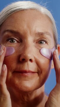 An elderly woman with patches under her eyes looks at the camera on a monochrome studio background, vertical video