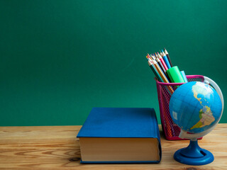 A book, a metal cup with pencils and a globe on the table against the background of a green board.