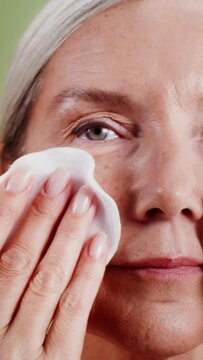 An elderly gray-haired model wipes her face with cotton pads, close-up. Vertical video, plain background