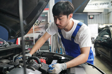 Male car operator wearing blue overalls,cap and gloves working under the hood of white car and checking attentively serviceability of engine at repair garage. Concept of car maintenance