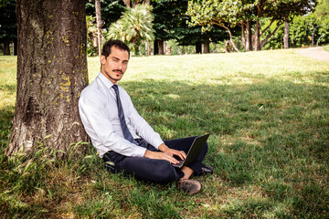 Obraz premium The beautiful businessman is working in a park. The young businessman is sitting on the lawn and leaning against a tree while he writes a work email.
