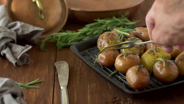 mini potatoes with spices and rosemary on the grill. On a brown wooden table. A man breaks a potato, pins it on a fork and eats it