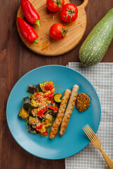 A plate of vegetables with grilled sausages on the table next to tomatoes, eggplant on planks and a wooden fork.