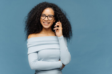 Indoor shot of positive lovely woman has telephone conversation, keeps mobile phone near ear, focused aside, wears casual jumper, isolated on blue background. People, technology, communication