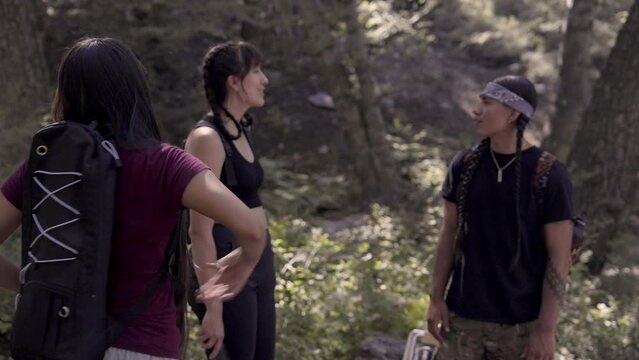 Young Native American hikers with backpacks talk under oaks and pines in the forest.