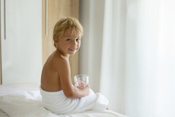 Cute blond toddler child with white tower around belly, sitting in bed with glass of water after bath