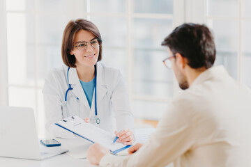 Young female doctor consults patient, shows information and prescription in clipboard, talk about medical checkup, pose at clinic, explains health insurance contract, read service agreement.