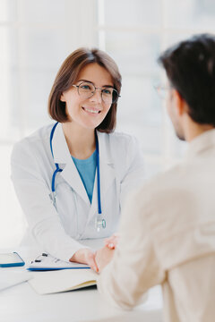 Medical trust concept. Vertical image of female doctor reassures her male patient, hold hand and talk calmly about illness cure, gives professional consultation and support, pose at doctors office
