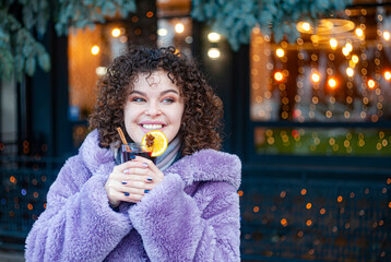 Smiling curly haired woman in trendy violet coat drinks mulled wine outdoors 