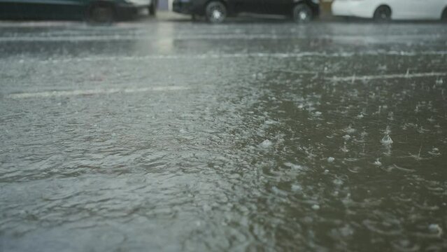 Close-up of raindrops fall on the road with cars driving in the background. Heavy torrential rain in the city.