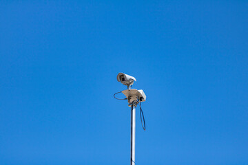 A CCTV security camera on top of a light pole with blue sky behind it.