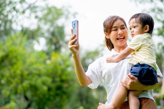 Young Asian mother hold little boy and use mobile phone for selfie photo shoot in garden and they look happy with holiday time in the morning together.