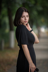 Portrait of a young beautiful dark-haired girl in a black dress in the park.