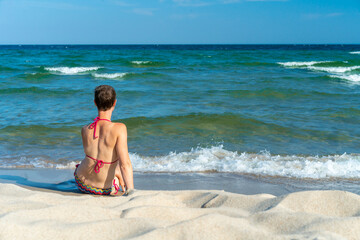 Fototapeta na wymiar Attractive sporty short haired woman in her thirties, wearing bikini, sitting on a beach close to the sea, looking at the horizon. Concept for holiday, vacation, relaxation, health, joy, recreation.