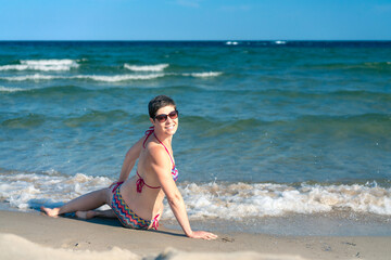 Attractive sporty short haired woman in her thirties, wearing bikini, sitting on a beach close to the sea, looking back over her shoulder. Concept for holiday, vacation, relaxation, health, joy.