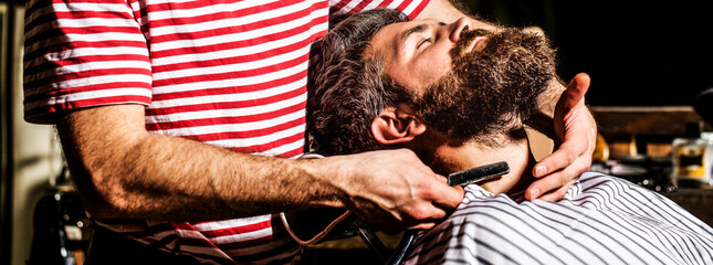 Men barber shaves his beard. Bearded male sitting in an armchair in a barber shop while hairdresser...
