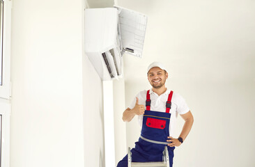 Happy young man in uniform standing on ladder under new air conditioner unit in white room, smiling...