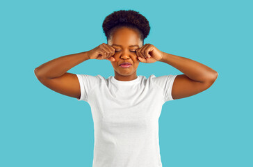 Fake tragedy. Funny capricious young african american woman grimacing and pretending to cry against light blue background. Girl in white t-shirt wipes her eyes from tears with cranky smile on her face