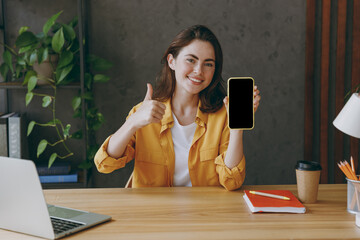 Young employee business woman wear casual yellow shirt hold in hand use mobile cell phone blank...