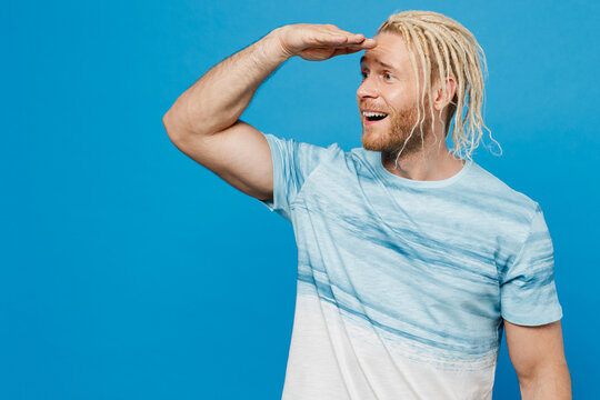Young fun blond man with dreadlocks 20s he wear white t-shirt look camera hold hand at forehead look far away distance isolated on plain pastel light blue background studio. People lifestyle concept.