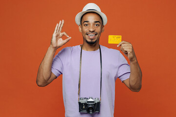 Traveler black man wear purple t-shirt hat hold credit bank card show ok isolated on plain orange...