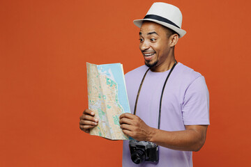 Traveler black shocked man wear purple t-shirt hat hold read map isolated on plain orange color background. Tourist travel abroad on weekends spare time rest getaway. Air flight trip journey concept.