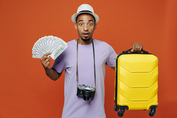 Traveler black man wear purple t-shirt hat hold suitcase fan cash money isolated on plain orange color background Tourist travel abroad on weekends spare time getaway Air flight trip journey concept.