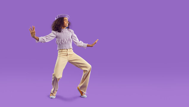Narrow shot of overjoyed teen African American girl in casual clothes have fun dancing on purple studio background. Happy biracial teenager make funny dancer moves. Copy space.