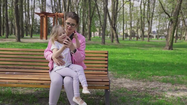 A caregiver and a girl are sitting on a park bench and are busy on a smartphone.