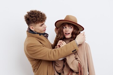 a cute couple is standing on a white background, wearing a coat. The man gently touches the ends of the woman's hat while she looks away in embarrassment