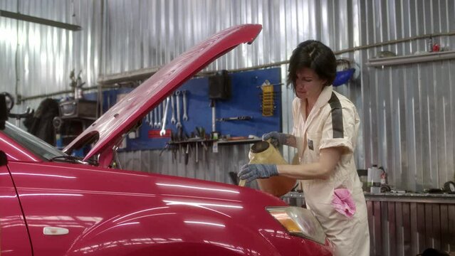 woman mechanic in light beige overalls pours oil into car from special watering can in car service. brunette is servicing red passenger car at scheduled maintenance in garage.