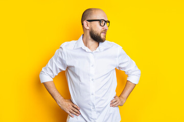 Young bearded man in glasses looks away holding his hands on his waist on a yellow background