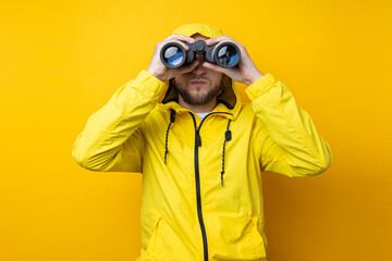 Young man in yellow raincoat looking through binoculars on yellow background