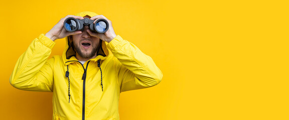 Fototapeta premium Surprised young man in yellow raincoat looking through binoculars on yellow background. Banner