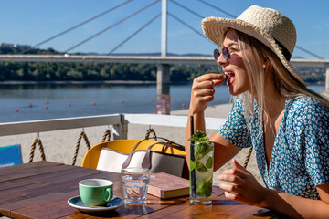 A beautiful young woman wearing a straw hat and sunglasses, having a cocktail and enjoying a lovely summer afternoon in a beach cafe by the river.