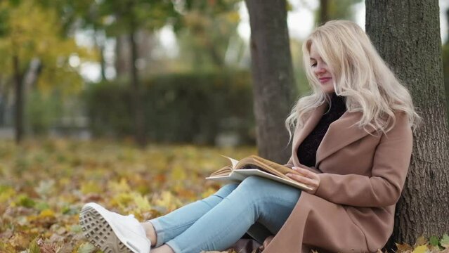 Romantic fall. Happy woman. Enjoying reading. Beautiful smiling lady sitting ground under tree in park with interesting book slow motion.